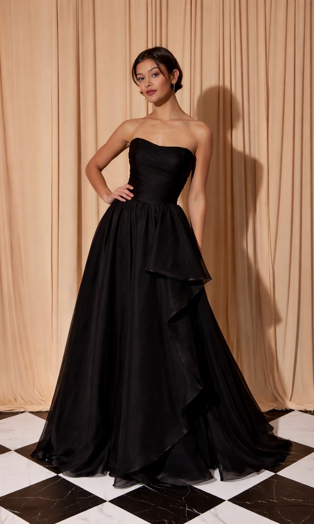 A woman in the Eureka EK206 long prom dress in black stands on a black and white checkered floor before beige curtains, her strapless organza gown radiating sophisticated elegance.